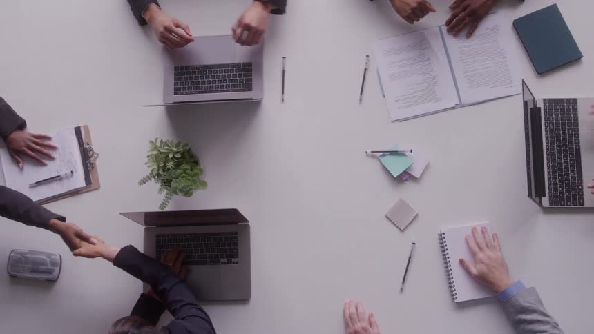 People sitting at a table typing on laptop computers and reaching over to shake hands