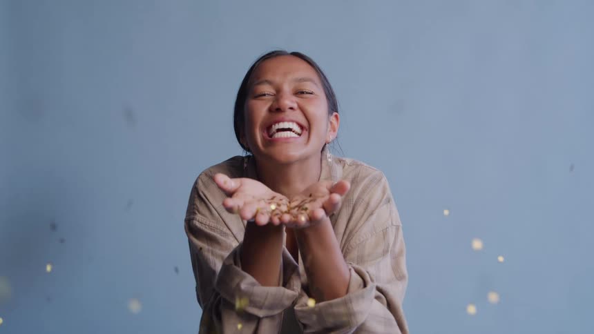 A joyful woman smiles as she blows a handful of golden glitter towards the camera