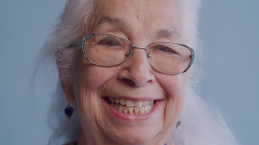 A smiling happy elderly female with long white hair and wearing eyeglasses is shown from the neck up