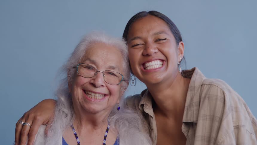 A younger woman with her arm around an older woman turns from smiling at each other to smile brightly together for the camera against a plain blue background