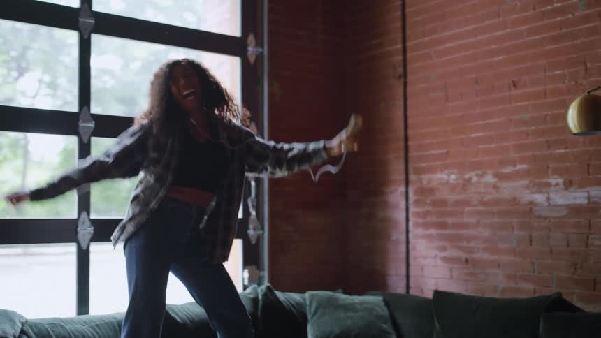 A young woman with long curly hair stands on a couch in her living room at midday excitedly dancing while wearing wired earbuds