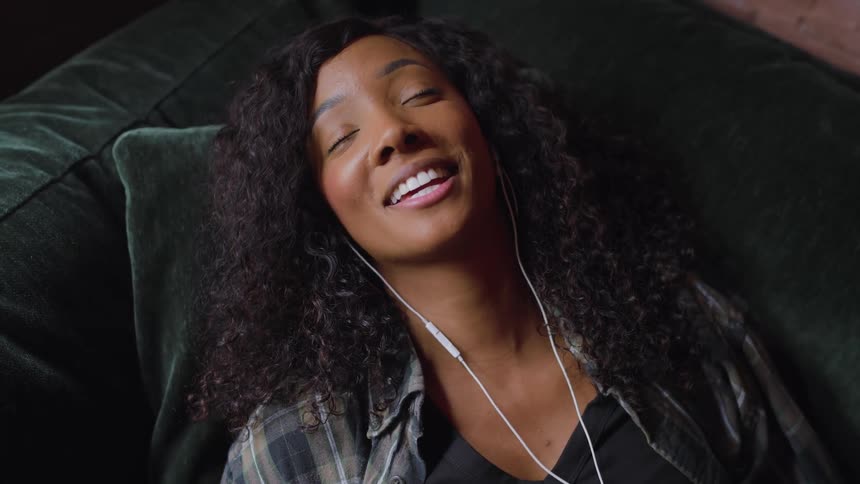 A black female wearing ear buds is smiling and singing as she lays back on a couch