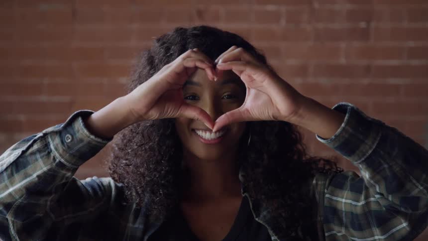 Woman smiles while forming a heart shape with her hands in front of a brick wall