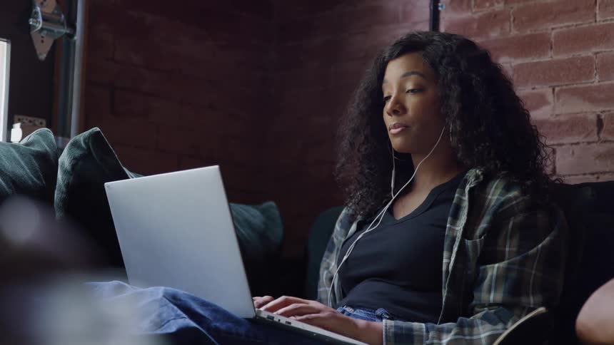 Woman uses a laptop while listening through earphones seated on the couch