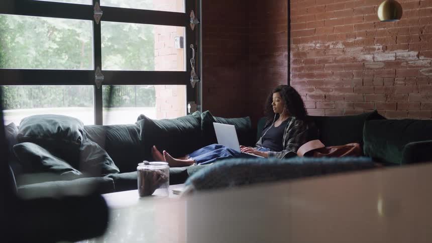 A woman relaxes on a large couch in a modern loft working on her laptop with earphones