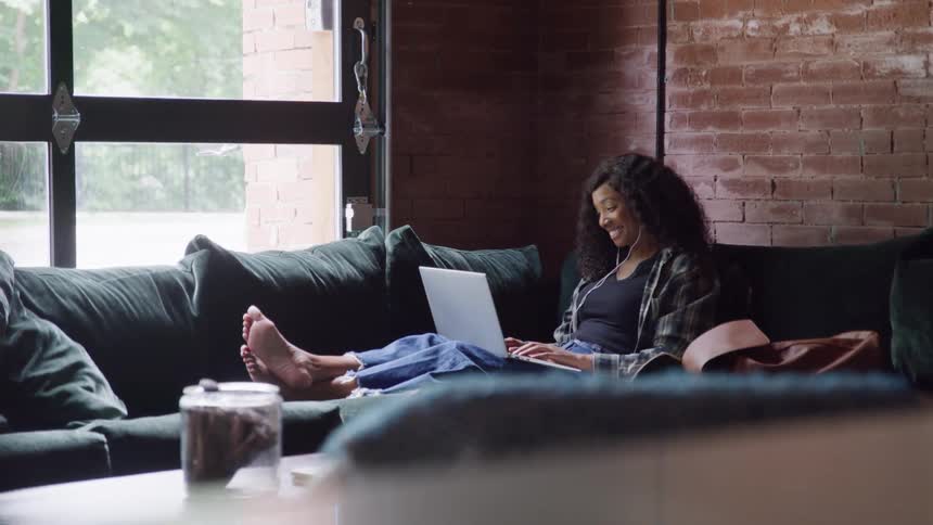 A woman sits on her sofa while waving at her laptop screen
