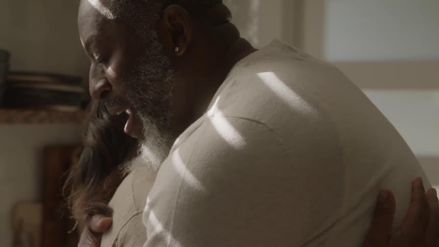 An older grey bearded man and a woman share a tender affectionate hug in a softly lit room