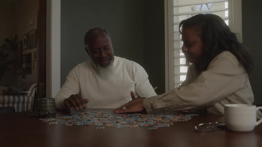 An older black couple are happy and smiling as they sit at a dining room table solving a jigsaw puzzle