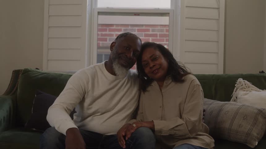 Couple sitting on the living room couch look into each others eyes and smile as they turn their heads