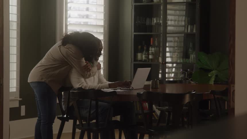 A middleaged black man sits at the dining table typing on his laptop while a woman walks up behind him and gives him a hug at midday in their home