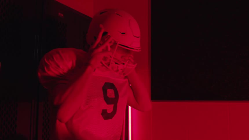 A football player puts on his helmet in a locker room and stands up