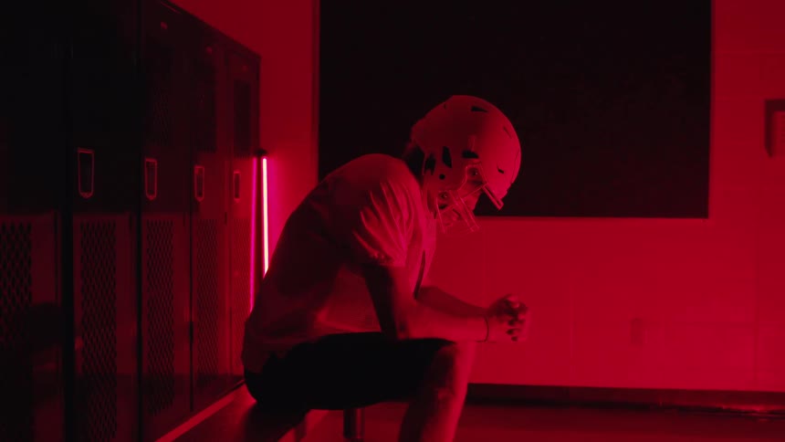 Football player sits on a bench in a locker room illuminated by red lighting