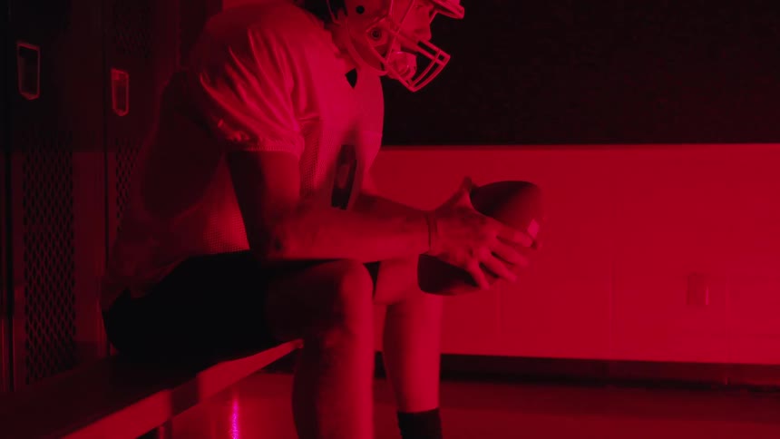 Male football player holds a football while sitting on a bench in a locker room lit by red lighting