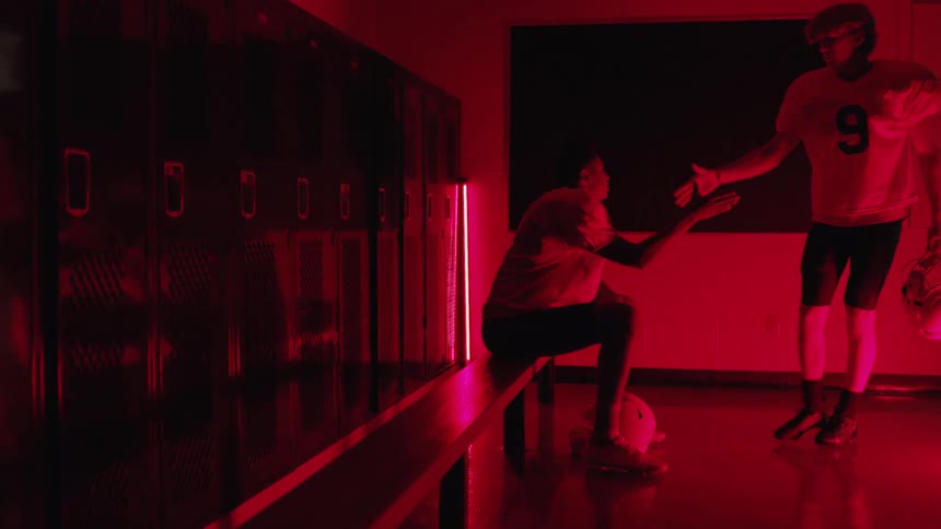 An american football player sits alone in the locker room looking sad then another player walks in gives him a handshake and walks out