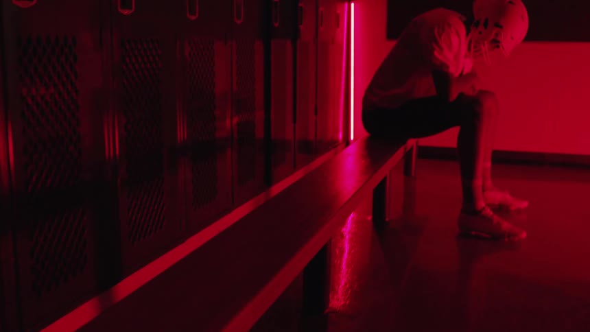Young man wearing a football uniform sits on a bench in the locker room filled with red light