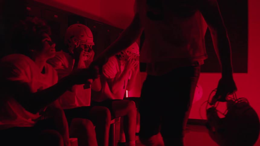 Football players gather in a locker room lit by red lighting