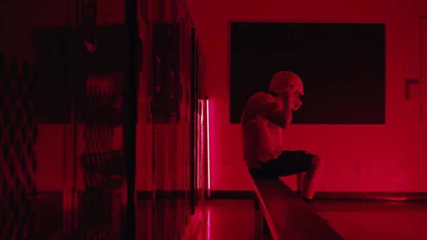 A young male football player wearing a light shirt and shorts sitting on a wooden bench in a long hallway lined with dark lockers dramatically illuminated by harsh red and magenta light as he pulls on his protective football helmet