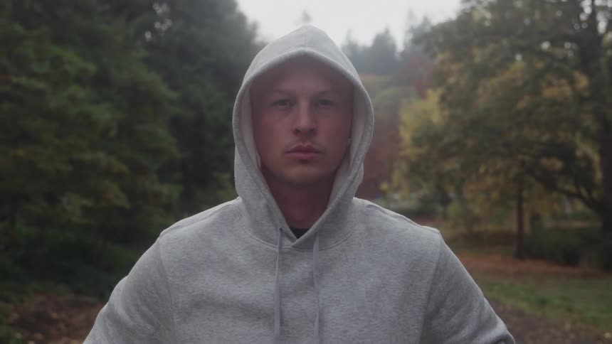 A young man wearing a hoodie standing outdoors at midday surrounded by trees