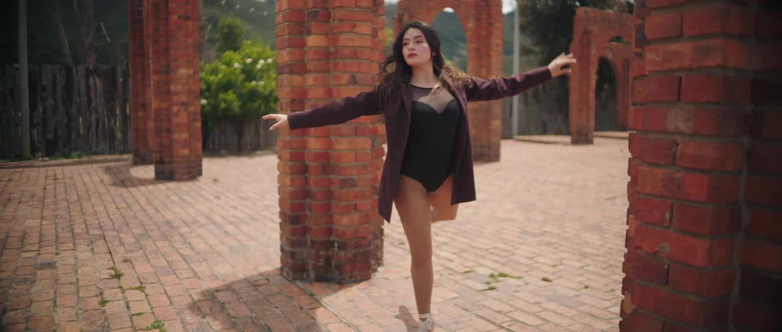 The video shows a young woman performing ballet in an outdoor setting characterized by red brick columns and archesshe is dressed in a dark leotard a sheer black top underneath and a dark blazer while wearing white pointe shoes