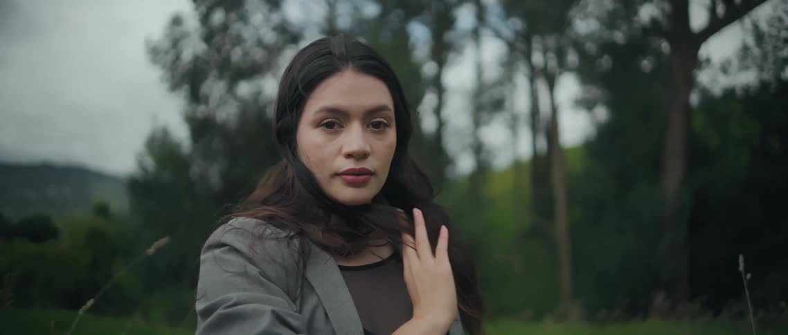 A woman stands in a forest clearing looking toward the camera on a bright day