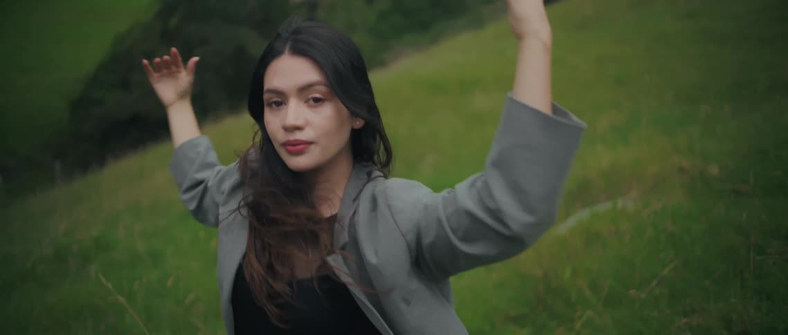 The video shows the woman dancing with stylized arm and hand movements in an expansive lush green fieldshe faces the camera initially then turns to the side maintaining a strong expressive posture while wearing a gray blazer and dark leotardtop