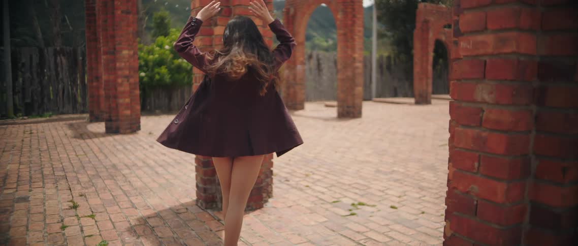 Young woman wearing a purple jacket and ballet shoes dancing surrounded by brick arches with trees and a hill in the background outdoors during day time