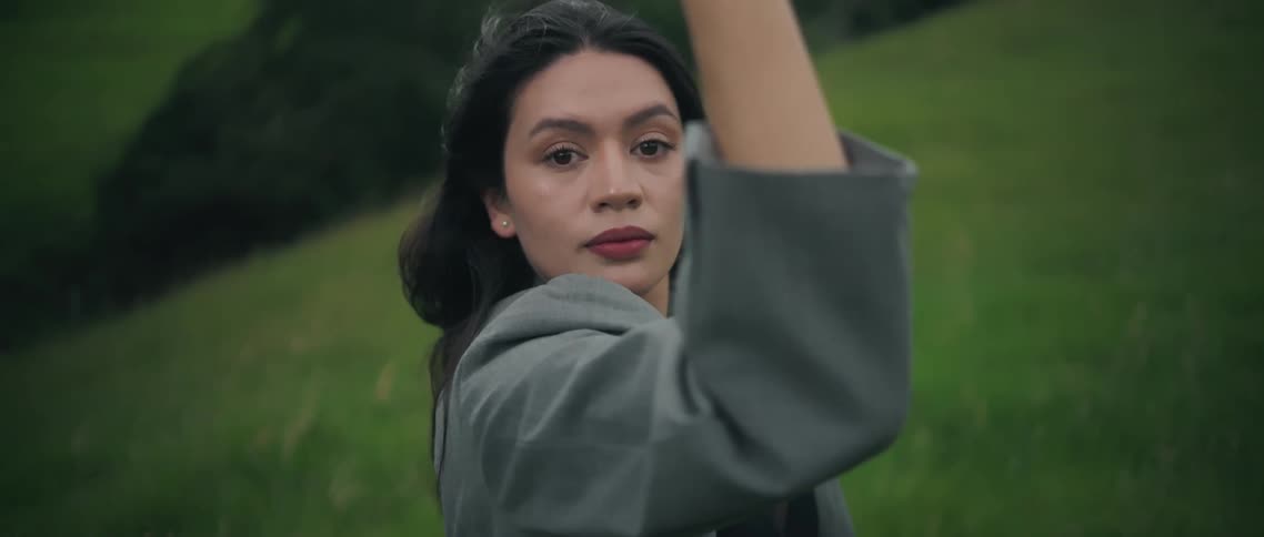 Woman standing on a grass covered hill holding up her arms in tai chi exercise as she looks into the camera