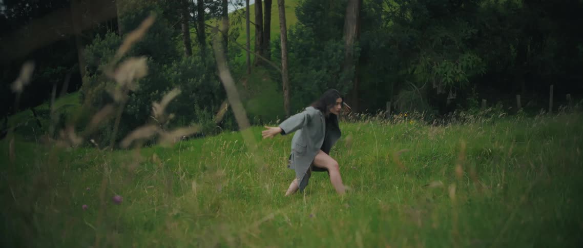 A young woman with long dark hair wearing an open grey jacket and black undergarments that expose her legs dances gracefully in agreen grassy field bordered by a dense forest