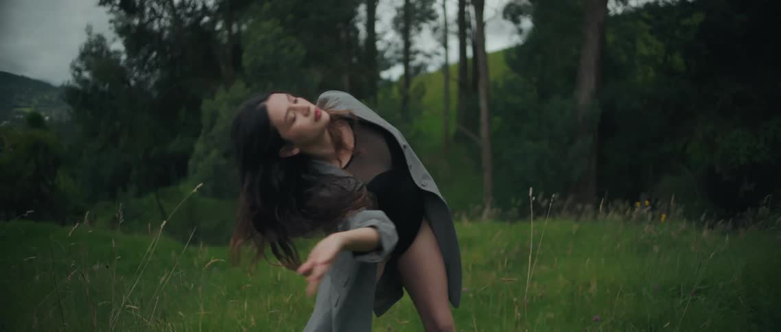A young woman in a flowing coat performs an expressive modern dance in a lush green meadow