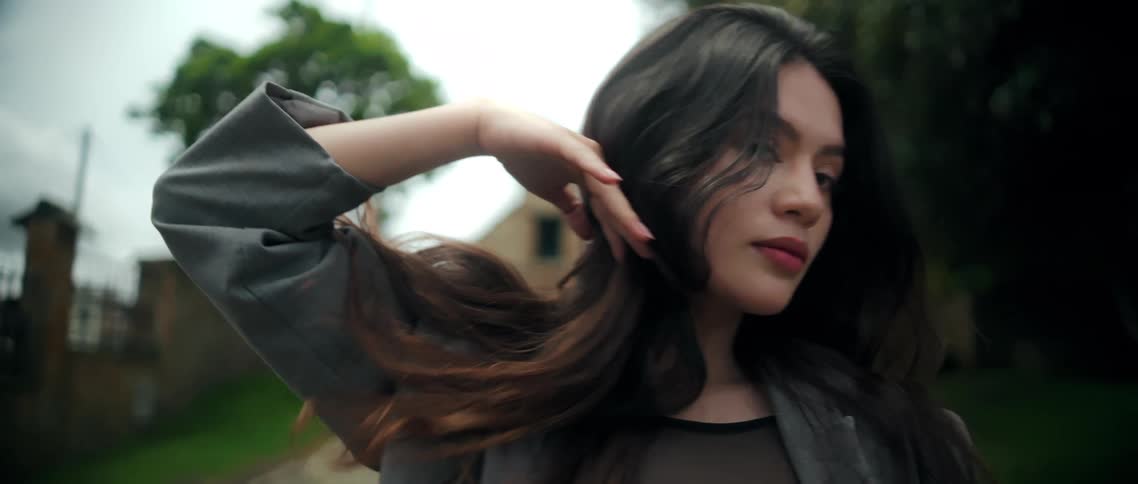 A woman with long hair is filmed outdoors initially leaning her head back with her eyes closed then bringing her gaze forward while using her hand to sweep her hair back behind her shoulder against a softly blurred backdrop of trees and an old structure
