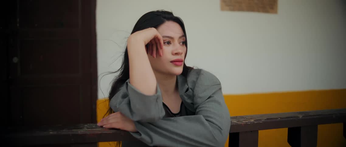 A woman leans on a rustic wooden railing gazing to the side against a white and yellow wall