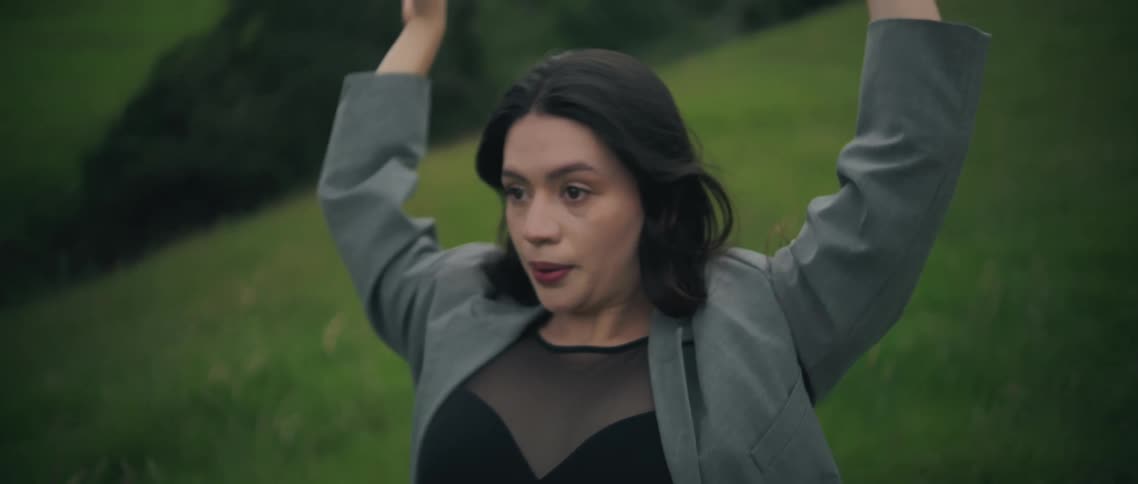 This video focuses on the same woman from the previous clip dancing expressively in a grassy fieldshe performs fluid movements with her arms and hair looking around intensely while wearing a gray blazer over a dark top