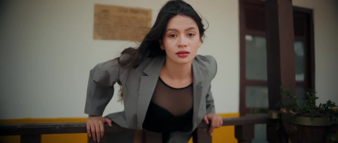 A young woman with long dark hair wearing a grey jacket over a black mesh top leans forward over a dark wooden railing looking intently forward against a background of a white and yellow building