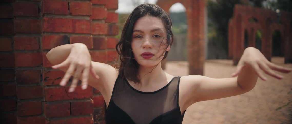 A woman performs slow dance movements beside a textured brick wall outdoors