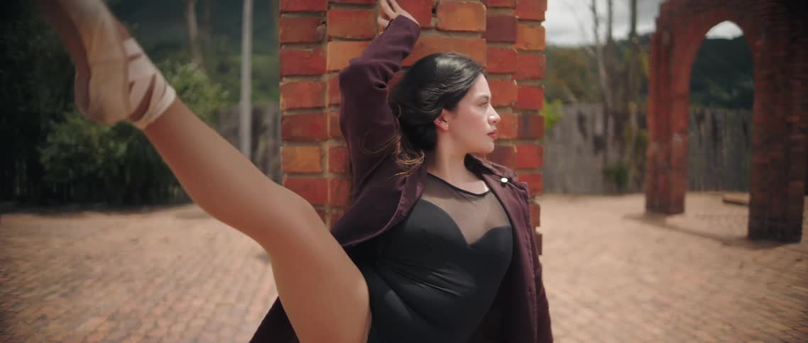 A tight shot focuses on a woman dressed in a black bodysuit and a maroon coat using a brick column as a prop for a dance pose she is captured holding a high extension leaning into the column with one arm raised while lifting her leg toward the camera w