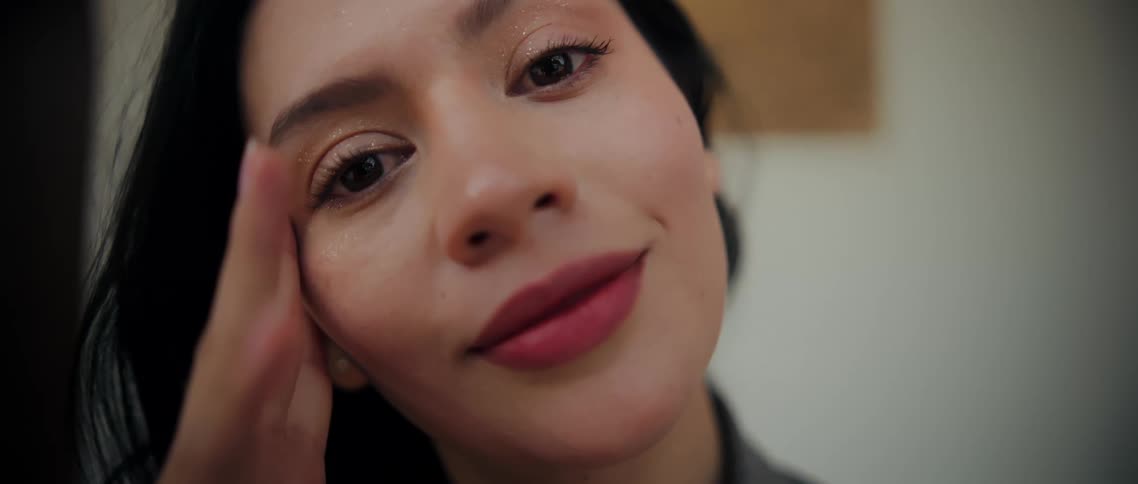 A womans face highlighting her glittering eye makeup and pinktoned lipstick shows her holding a subtle gentle smile while her fingers rest lightly against the side of her forehead