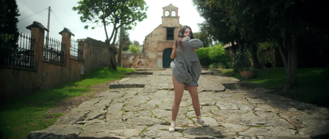 A woman in a black bodysuit light grey coat and ballet shoes dances on a cobbled stone path leading up a slight incline she performs a dynamic routine including spinning with her coat flaring out and striking ballet poses with her arms raised all fra