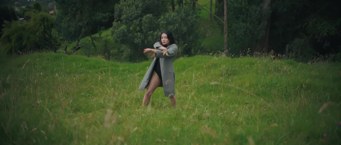 A woman wearing a dark bodysuit and a long grey coat performs a slow expressive dance in a grassy field she moves fluidly raising and lowering her arms above her head while surrounded by tall green grass and trees in the background