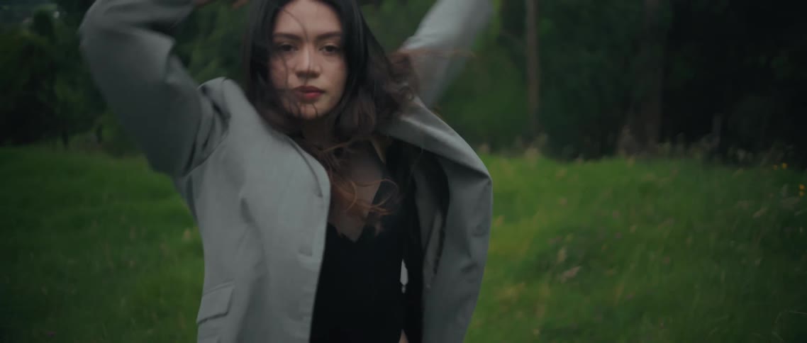 A young woman stands on a grassland at midday wearing a long unbuttoned coat over a black swimsuit gracefully performing a dance move