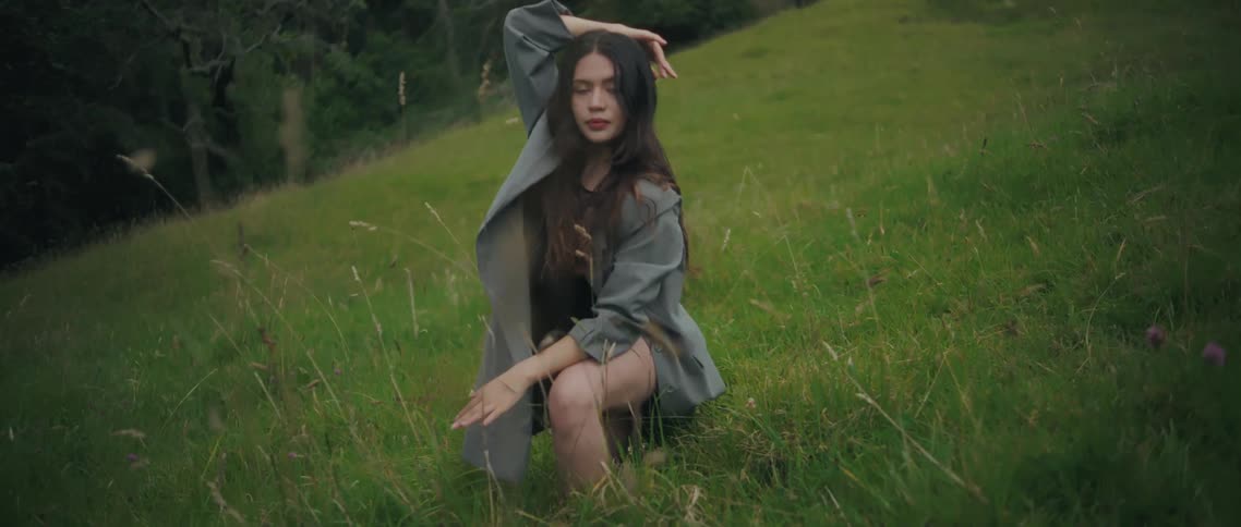 Woman kneels on the sloped grass while resting one hand on her head and holding an elegant posture