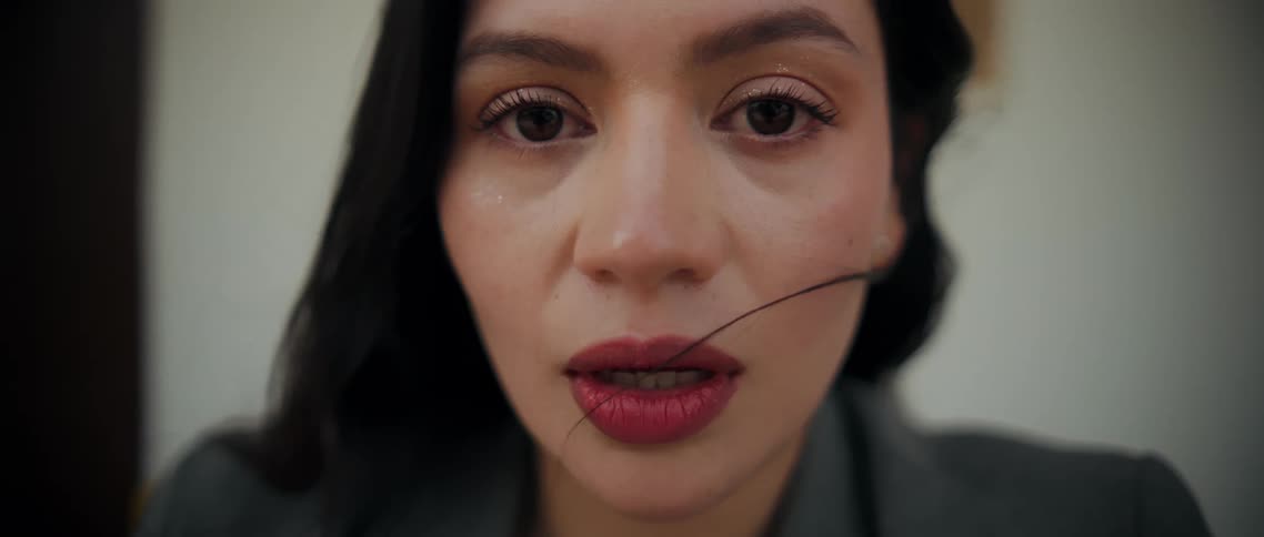 A young woman with dark hair and striking makeup wearing a grey jacket looks intensely forward in a closeup shot that captures a thoughtful or serious mood