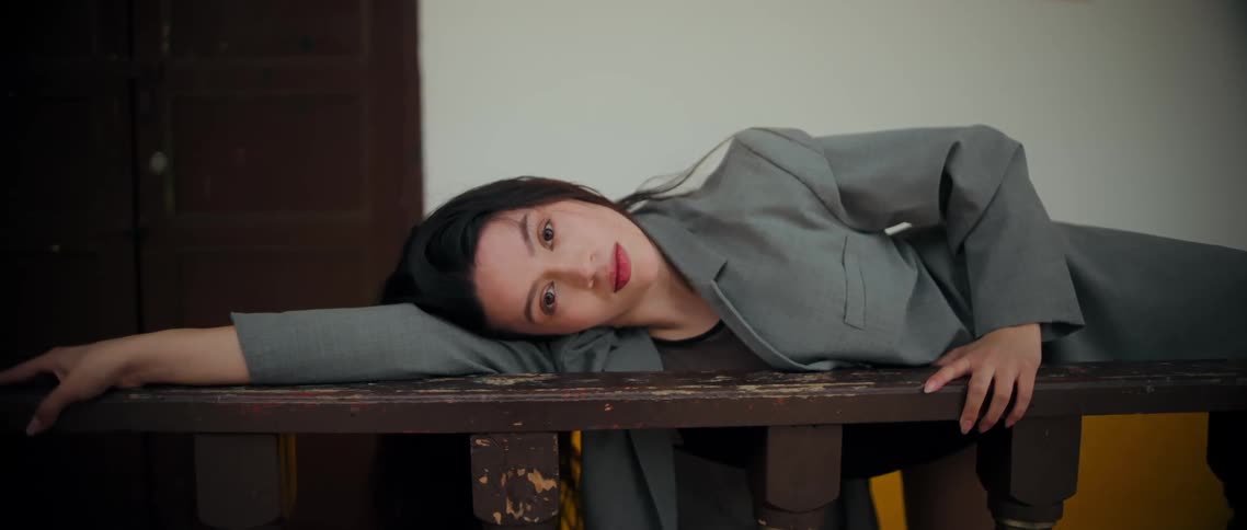 A woman lies on a wooden indoor railing while looking directly at the camera