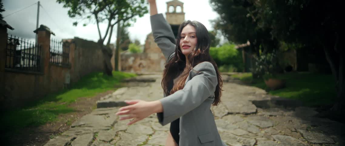 A woman dances with raised arms on a cobblestone path in front of an old stone building