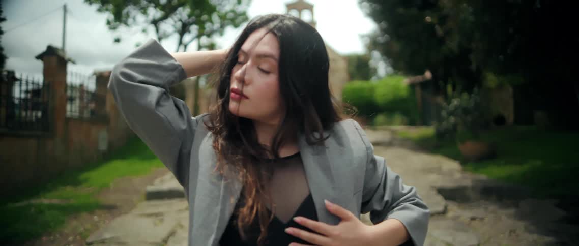 A young woman with long brown hair dressed in an ashcolored suit performs a dance move beside a building fence outdoors at midday