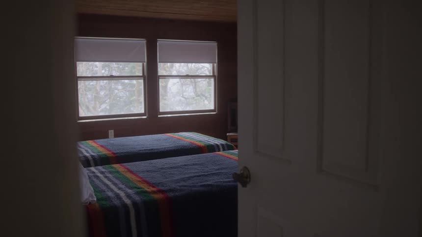 The camera slowly moves forward through a white doorway revealing a rustic woodpaneled bedroom with two twin beds sitting under bright windows