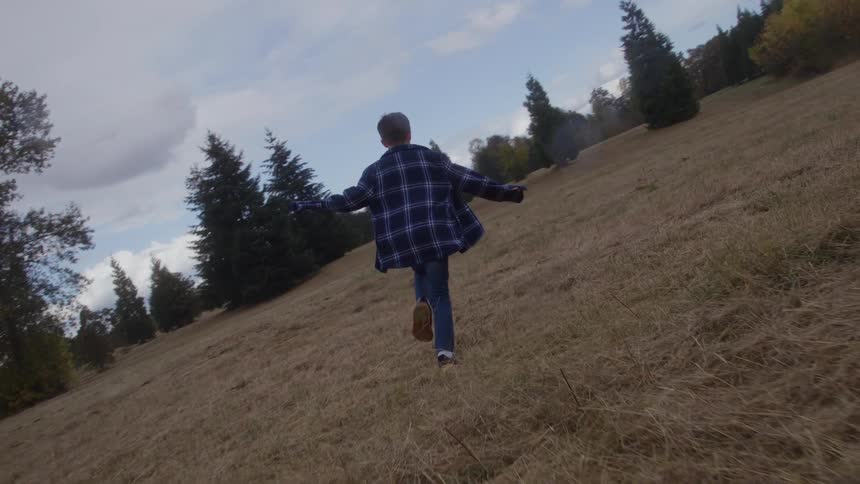A person wearing a blue plaid jacket runs up a steep dry grassy hillside with their arms extended out to the sides the hillside is bordered by evergreen trees and the sky is bright with some scattered clouds overhead