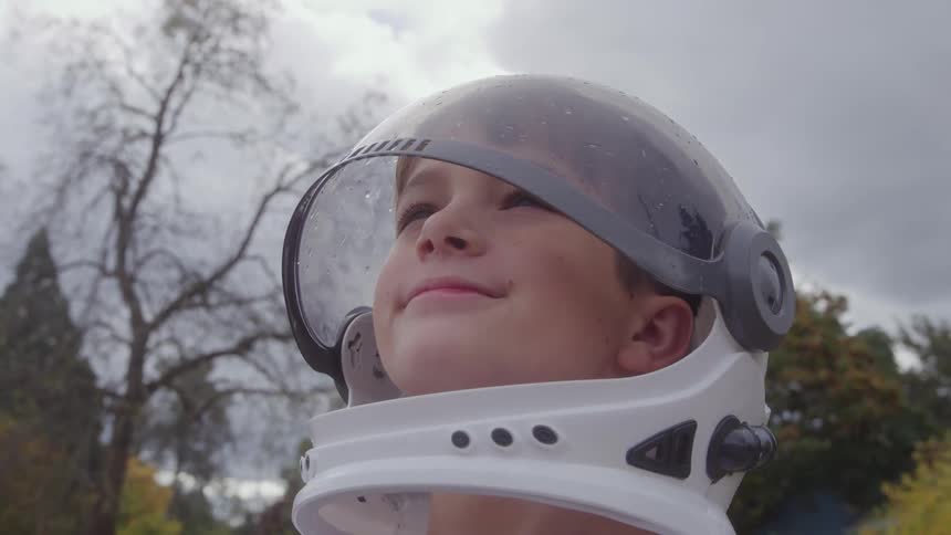 A young boy in an astronaut helmet looks up at the sky with a hopeful smile