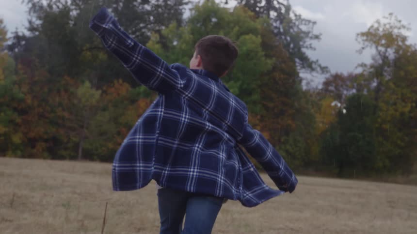 A young person wearing a blue plaid jacket and jeans runs across a wide dry field with their arms outstretched the background shows a line of trees displaying a mix of green yellow and reddish autumn foliage under a cloudy sky