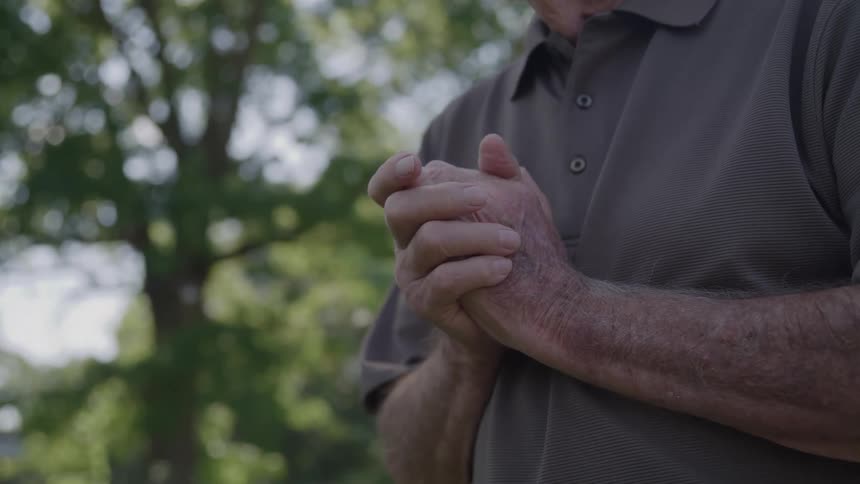 Elderly persons hands clasped in front of body outdoors with sunlit trees in the background