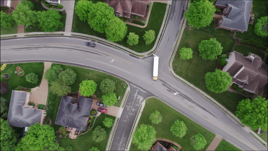 An aerial view captures a quiet suburban neighborhood with large houses lush green lawns and trees where a white semitruck is driving through a winding asphalt street intersection